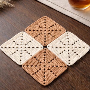 May include: Four handmade crochet coasters in cream and tan. Each square coaster features a starburst design with openwork details. The coasters are arranged on a dark wood surface, suggesting a cozy, handcrafted aesthetic.