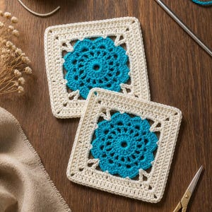 May include: Two square crochet motifs featuring a cream-coloured border and a vibrant turquoise floral centre. The pieces are crafted with intricate yarn stitches, showcasing a geometric design that combines a circular inner pattern with a square frame.
