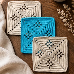 May include: Three square crochet coasters in beige and turquoise blue, with a floral design. The coasters are placed on a wooden surface, with a cup of coffee and a beige cloth in the background. The coasters are approximately 4 inches square.