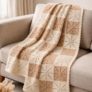 Op de afbeelding: Een handgemaakte gehaakte deken in een neutraal kleurenpalet van crème en bruin, gedrapeerd over een beige bank. De deken heeft een geometrisch vierkant patroon met een geschulpte rand, wat een gezellig en decoratief huistextiel creëert.