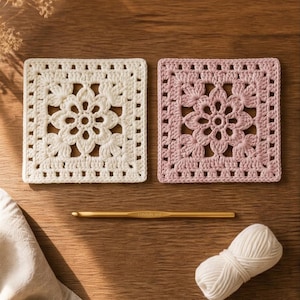 May include: Two square crochet coasters, one cream and one pink, with a floral design. A gold crochet hook and a ball of white yarn are also present. The coasters are on a wooden surface, suggesting a handmade craft theme.