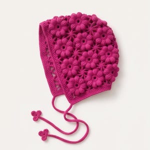 Puede incluir: Un gorro de ganchillo magenta con un diseño floral. El gorro presenta flores en relieve en la parte superior y los lados. Tiene lazos con extremos en forma de flor. El gorro está hecho de un hilo suave y texturizado.