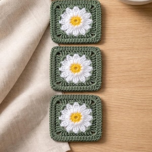 May include: Three square crochet coasters with a daisy design. Each coaster features a white daisy with a yellow centre, set against a green square background. The coasters are handmade and placed on a wooden surface.