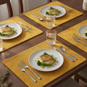 May include: A set of rectangular mustard yellow crocheted placemats arranged on a wooden dining table. Each placemat features a textured, woven pattern and supports a white ceramic plate with a meal, a clear glass, and stainless steel cutlery.