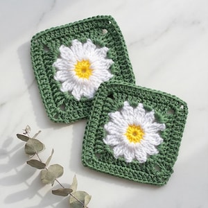 May include: Two square crochet coasters. Each coaster features a white daisy with a yellow centre, set against a green background. The coasters are handmade and placed on a white marble surface.