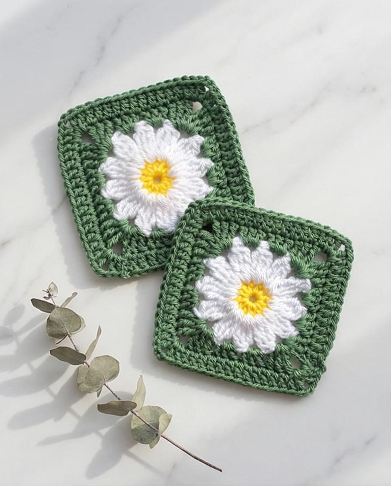 May include: Two square crochet coasters. Each coaster features a white daisy with a yellow centre, set against a green background. The coasters are handmade and placed on a white marble surface.