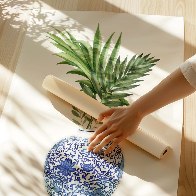 K&ouml;nnte beinhalten: Eine wei&szlig;e Keramikvase mit einem blauen und wei&szlig;en Blumenmuster wird in eine wei&szlig;e Papierrolle gerollt. Die Vase steht auf einer wei&szlig;en Oberfl&auml;che mit einer gr&uuml;nen, bl&auml;ttrigen Pflanze im Hintergrund.
