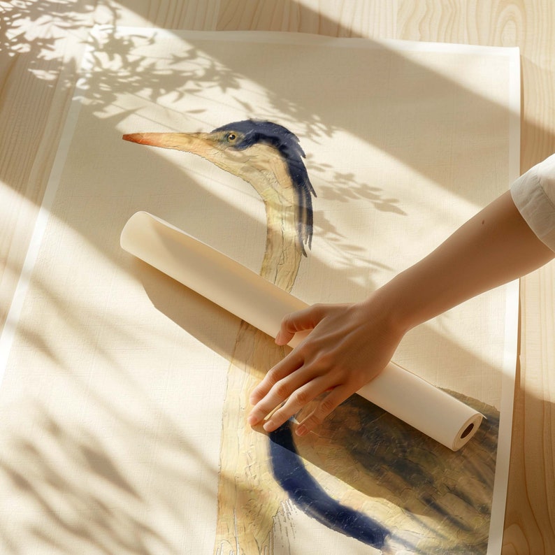 May include: A hand rolls up a white canvas print featuring a detailed illustration of a blue and yellow heron.