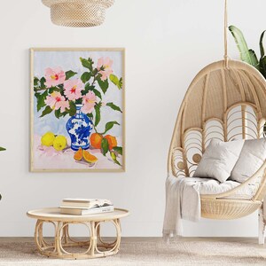 May include: A painting of pink flowers in a blue and white vase with oranges and lemons on a pink table. The painting is framed and hanging on a white wall.