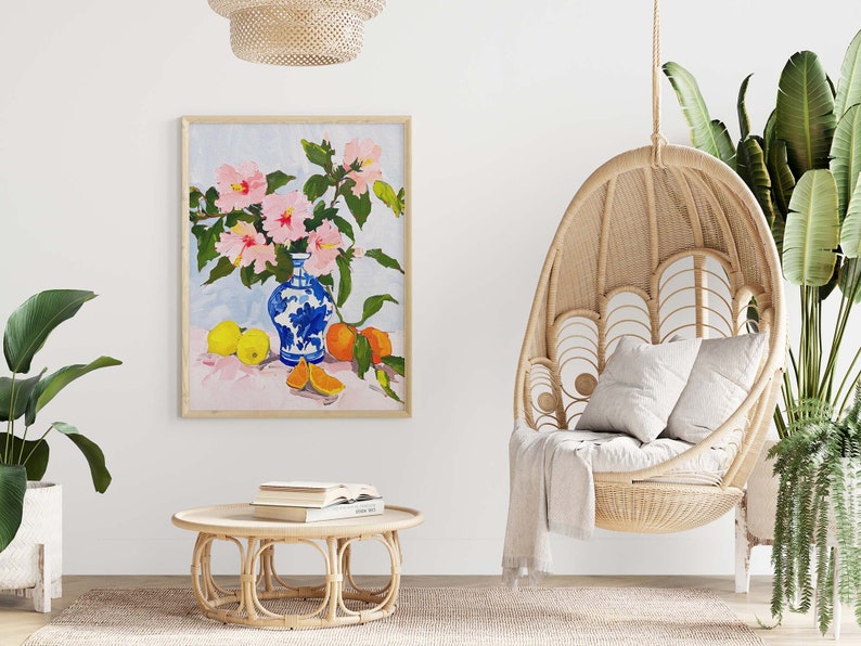 May include: A painting of pink flowers in a blue and white vase with oranges and lemons on a pink table. The painting is framed and hanging on a white wall.