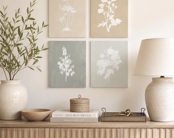 May include: Four square wall art prints featuring white botanical illustrations on beige and sage green backgrounds, arranged in a grid. The prints are displayed above a wooden sideboard with a lamp, a woven box, books, and a decorative tray.