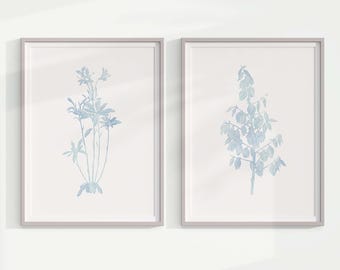 May include: Two framed botanical prints. Each print features a light blue watercolor illustration of a plant against a white background. The frames are a light gray color. The print on the left shows a cluster of flowers, and the print on the right shows a tree.