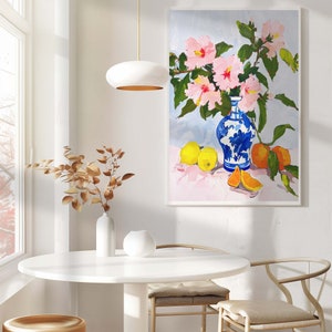 May include: A blue and white porcelain vase with pink hibiscus flowers and yellow oranges on a pink background. The vase is sitting on a table with a white lamp and a white vase with brown branches.