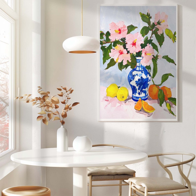 May include: A blue and white porcelain vase with pink hibiscus flowers and yellow oranges on a pink background. The vase is sitting on a table with a white lamp and a white vase with brown branches.