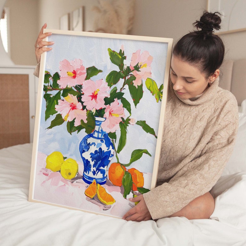 May include: A painting of pink hibiscus flowers in a blue and white porcelain vase with lemons and oranges on a pink background.
