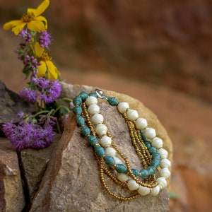 May include: A multi-strand necklace with white and green beads, and gold beads. The necklace is resting on a rock.