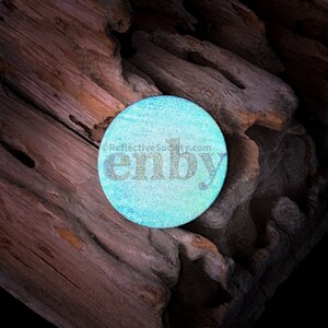 May include: A round, iridescent blue and green sticker with the word "emby" printed in black letters. The sticker is on a piece of weathered wood.