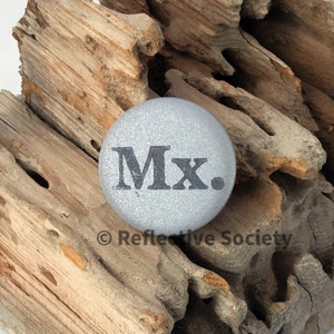 May include: A silver round button with the text "Mx." printed in black.