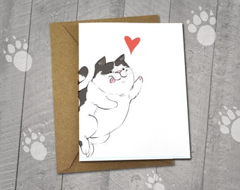 May include: A blank greeting card featuring an illustration of a happy, cartoon cat with black and white markings, a pink tongue, and a red heart. The card is paired with a brown envelope. The text on the card reads "Original Illustration 5x6.5" Blank Card / 5x7" Envelope".