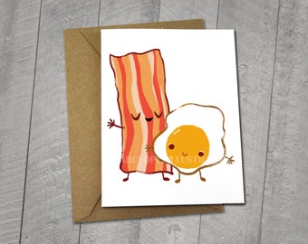 May include: A blank greeting card featuring an original illustration of a smiling cartoon bacon strip and a smiling cartoon fried egg. The card is 5x6.5 inches and comes with a 5x7 inch envelope.