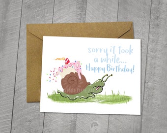 May include: A blank birthday card with a cartoon snail carrying a cupcake on its back. The card reads "sorry it took a while... Happy Birthday!" in light blue text. Includes a brown envelope. The card measures 5x6.5 inches.