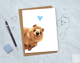May include: A greeting card featuring a brown quokka illustration with a light blue heart above its head. The card is placed on a light gray surface next to a mechanical pencil, a blue eraser, and a hand-drawn sketch of a quokka with hearts.