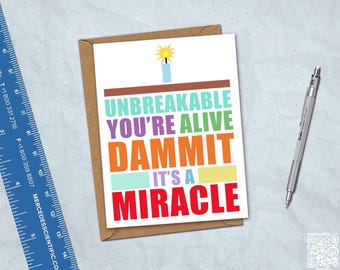 May include: A white greeting card with a brown envelope, featuring a blue candle with a yellow flame and the text: UNBREAKABLE YOU'RE ALIVE DAMMIT IT'S A MIRACLE in colorful block letters. A blue ruler showing inches and a silver mechanical pencil are visible.