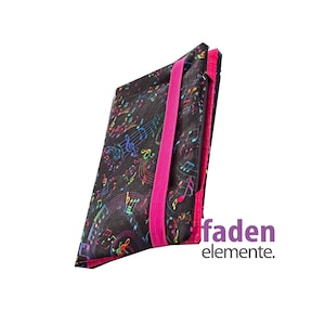 Könnte beinhalten: Eine schwarze Hülle mit einem bunten Notenmuster, gesichert durch ein leuchtend pinkfarbenes Gummiband. Der Text "faden elemente" ist sichtbar. Die Hülle dient zur Aufbewahrung.