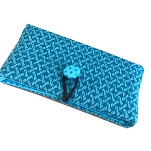 Puede incluir: Un estuche de tela turquesa con un patrón tejido y un cierre de botón azul con lunares. El estuche es rectangular y tiene un interior azul claro en contraste. Una banda elástica negra asegura el botón.