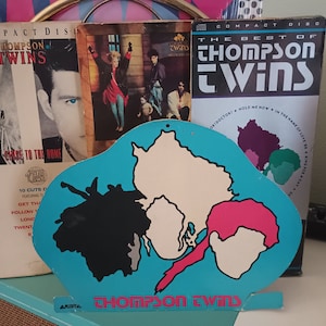 May include: A blue and white cardboard cutout of a map with the words "Thompson Twins" printed on the bottom. The map is shaped like a cloud and has three islands, one black, one white, and one pink.