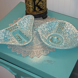 May include: Two asymmetrical ceramic dishes featuring a turquoise and white speckled finish. Each dish includes a row of small gold-colored circular indentations and a central recessed area with gold-colored accents on the interior surface.
