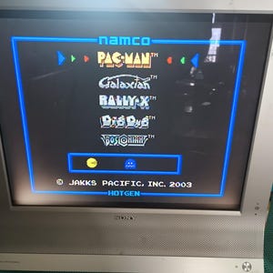 May include: A silver Sony television displays a video game menu with the names PAC-MAN, Galaxian, Rally-X, Dig Dug, and Bosconian. The screen has a blue border and the text "namco" at the top. The copyright reads "JAKKS PACIFIC, INC. 2003".