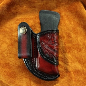 Może przedstawiać: Brązowa skórzana pochwa na nóż z kwiatowym wzorem i czarną skórzaną pętlą na pasek. Pochwa ma zatrzask i jest zaprojektowana do małego noża.