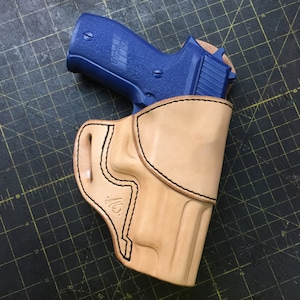Puede incluir: Una funda de cuero marrón con una pistola azul en su interior. La funda tiene un diseño cosido y un pequeño logotipo en el lateral.