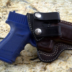 Può includere: Una pistola blu in una fondina di cuoio marrone con una cinghia di cuoio nero e bottoni a pressione argentati. La fondina ha cuciture bianche.