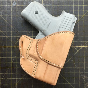 Può includere: Un fodero in pelle marrone con un design cucito, che contiene una pistola grigia.