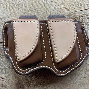 以下が含まれることがあります： 白のステッチが施された、薄い茶色の革の裏地が付いた、茶色の革製のダブルマガジンポーチです。