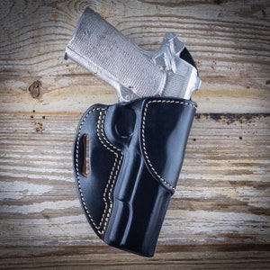 May include: A black leather holster with white stitching, holding a silver handgun. The holster has a curved design with a belt loop. The handgun is partially visible, showing its grip and slide. The background is a wooden surface.