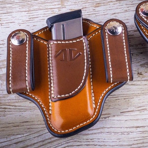 May include: A brown leather holster with a magazine holder and two belt loops. The holster has a stitched design and a black trim.
