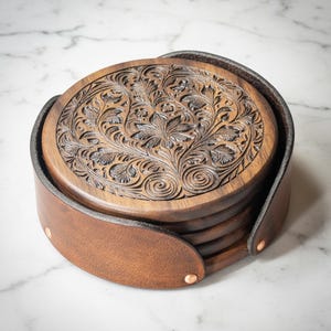May include: A stack of circular dark brown wooden coasters featuring intricate floral carvings on the top surface. The coasters are held together by a tan leather wrap secured with small metallic rivets along the curved exterior edge.
