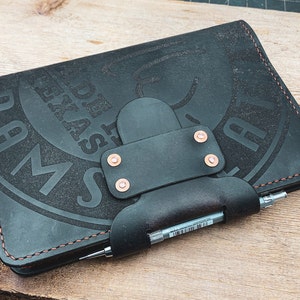 May include: A black leather journal with a pen holder and a stitched border. The cover features an embossed design with the text "MADE IN TEXAS" and "AUSTIN".