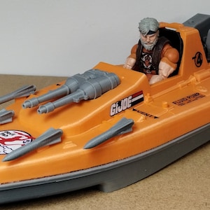 Pode incluir: Um barco de brinquedo laranja e cinza com o texto "G.I. JOE" e "FLUID PUMP" impresso na lateral. O barco tem projéteis cinza em forma de míssil e uma figura de ação sentada. O barco tem um emblema branco e vermelho na frente.
