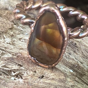 May include: A copper-toned ring with a teardrop-shaped, amber-colored stone. The ring's band has a twisted rope design. The stone is set in a copper bezel, creating a rustic and elegant look. The ring is displayed on a wooden surface.