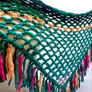 May include: A green crocheted shawl with colorful fringe. The shawl is made of a net-like material with a diamond pattern. The fringe is made of different colors of fabric, including orange, pink, and blue.