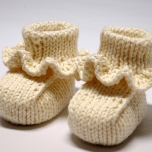 May include: A pair of knitted baby booties with a ruffled trim. The booties are cream colored and made of a soft yarn.