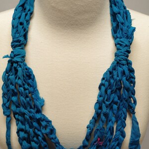 May include: A teal blue crocheted necklace made from fabric strips. The necklace is displayed on a white mannequin.