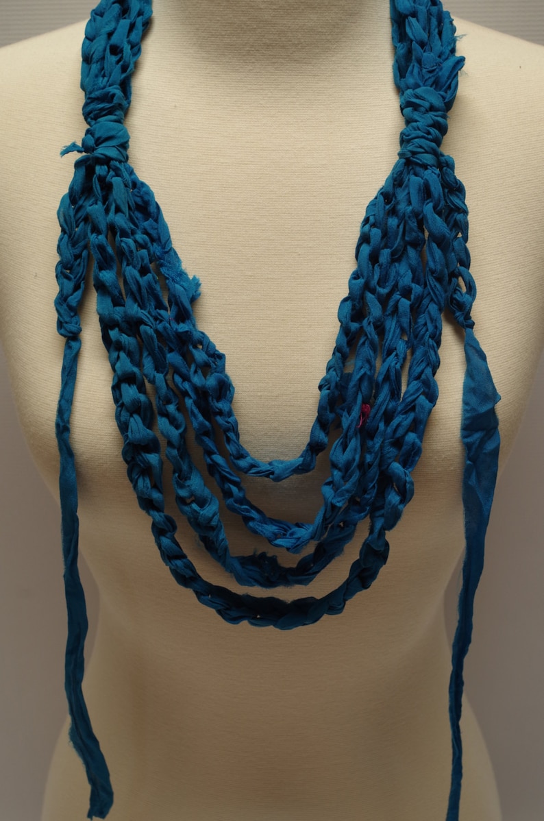 May include: A teal blue crocheted necklace made from fabric strips. The necklace is layered and has a long, flowing design.