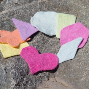 May include: A set of seven colorful knitted fabric scraps in various shapes, including a heart, a star, and a triangle. The fabric scraps are arranged in a circle on a gray rock.