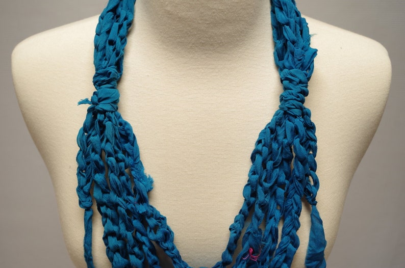 May include: A teal blue crocheted necklace made from fabric strips. The necklace is displayed on a white mannequin.