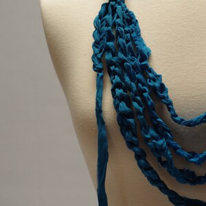 May include: A teal blue crocheted necklace with multiple strands of fabric. The necklace is displayed on a white mannequin.